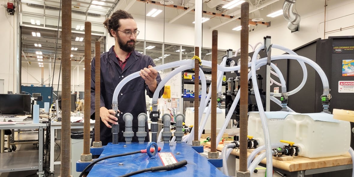 A man with glasses and a beard with dark hair is holding a white hose in a laboratory.