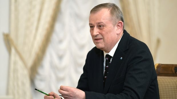 A man in a suit speaks at a table in an ornate room. 