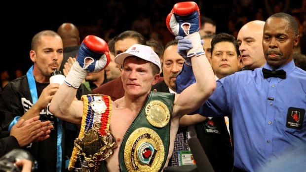A male boxer has his left arm held up by a referee in victory with two belts hanging over his shoulders.