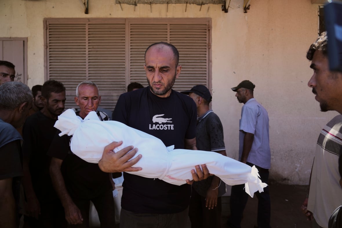 A man holds the body of a small child wrapped in a white substance.