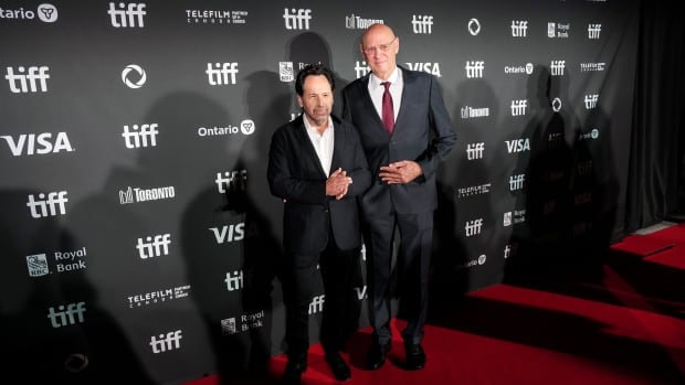 Two men in suits are photographed on a red carpet. 