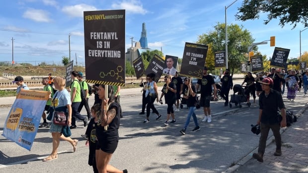 group of people walk down a city road holding black signs with text that read "Fentanyl is in everything" and "no thanks, I'm good."