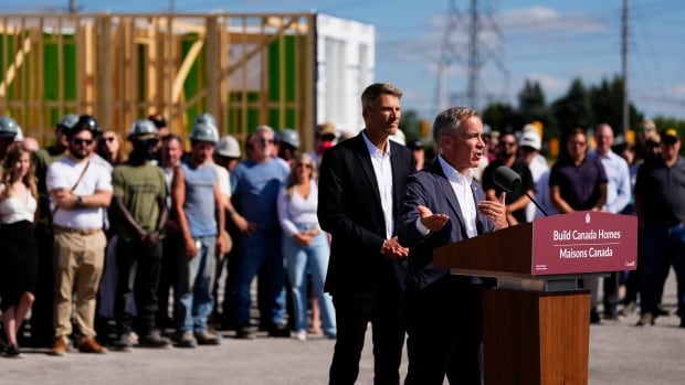 A man in a suit speaks at a podium as a crowd of construction workers watches in the background. Behind them is a house under construction.