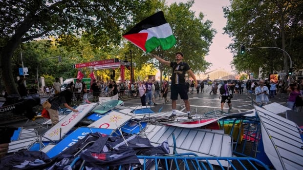 A protester waves a Palestinian flag on top of fences used as barricades to block the street to disrupt the twenty-first stage of the Spanish cycling race La Vuelta, from Alalpardo to Madrid, Spain, Sunday, Sept. 14, 2025. (AP Photo/Manu Fernández) Protesters wave flags while standing on a makeshift blockade made from steel barriers.