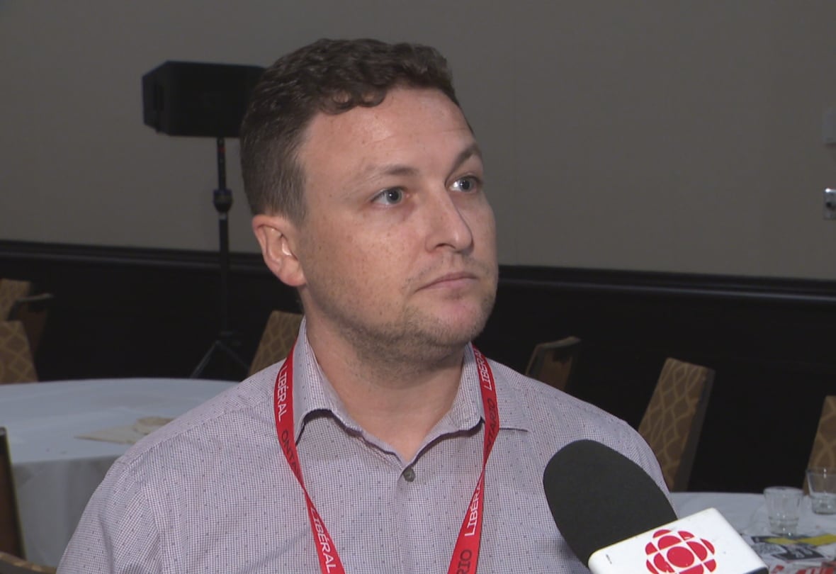 Man wearing a bright red Liberal lanyard speaks into a microphone.