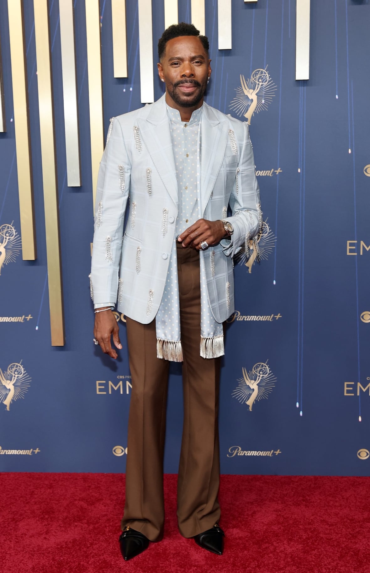 Ein Mann posiert für ein Foto in einem hellblauen Blazer und Hemd und einer braunen Hose.