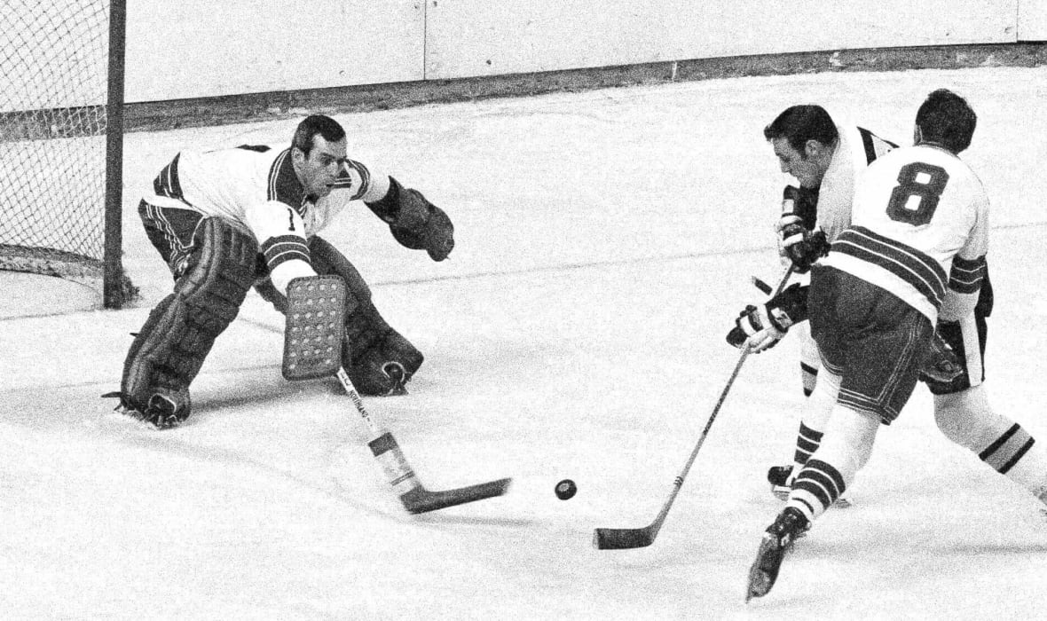A black and white photo of a hockey goalie in a white jersey extending his stick to poke a puck away. In front of him, two players jostle for positon.