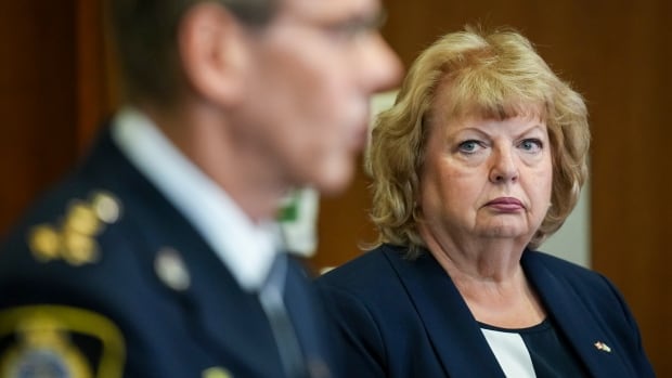 A woman with blond hair and a stern look on her face looks at a man in a police uniform as he speaks from a podium.