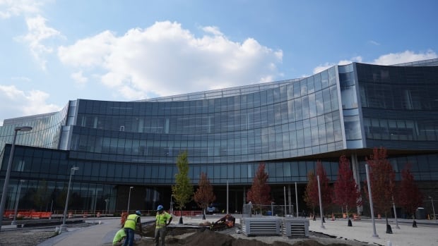 A large glass building curves inward in the middle. Trees dot the plaza in front of it and workers wear hardhats