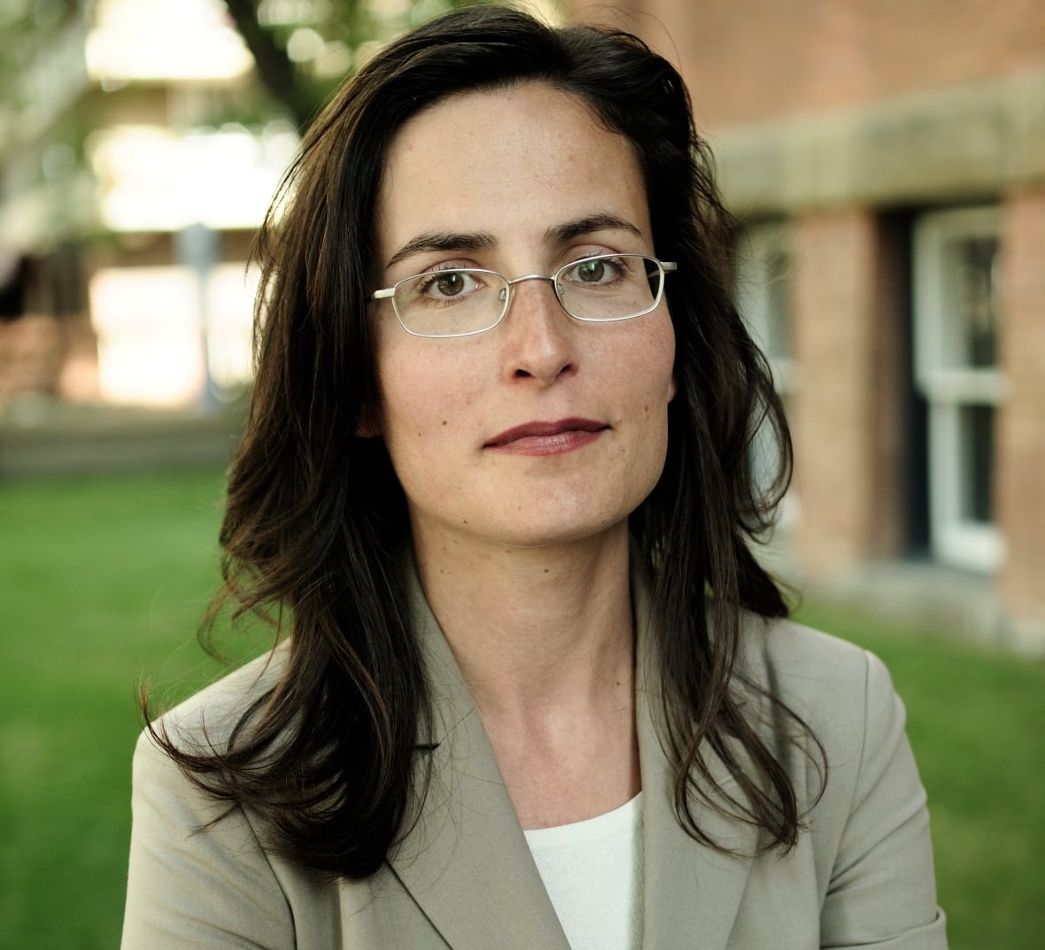 A woman wearing a grey suit stands outside, looking at the camera with her arms crossed over her chest.