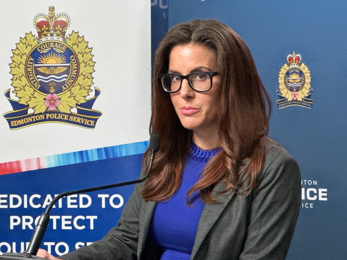 A woman with brown hair and glasses stands at a podium next to a logo for the Edmonton Police Service.