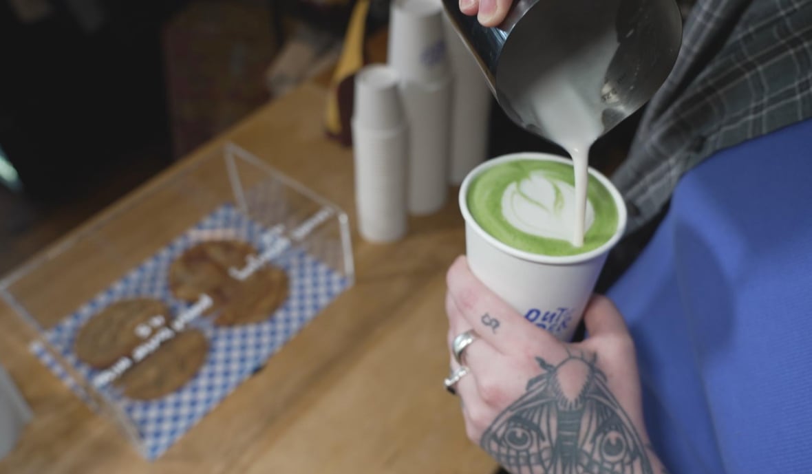 Tattooed hands hold a matcha latte in a paper cup and our a heart-shaped latte heart with milk. 