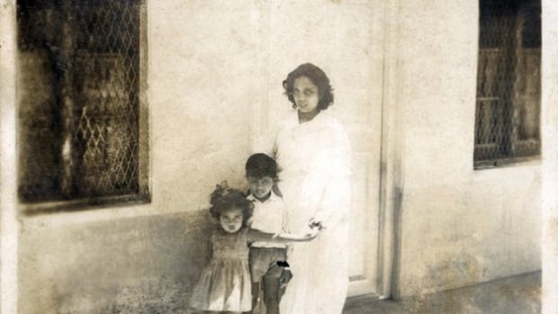 Two children and a woman stand in front of a house in an old sepia-tinted photo.