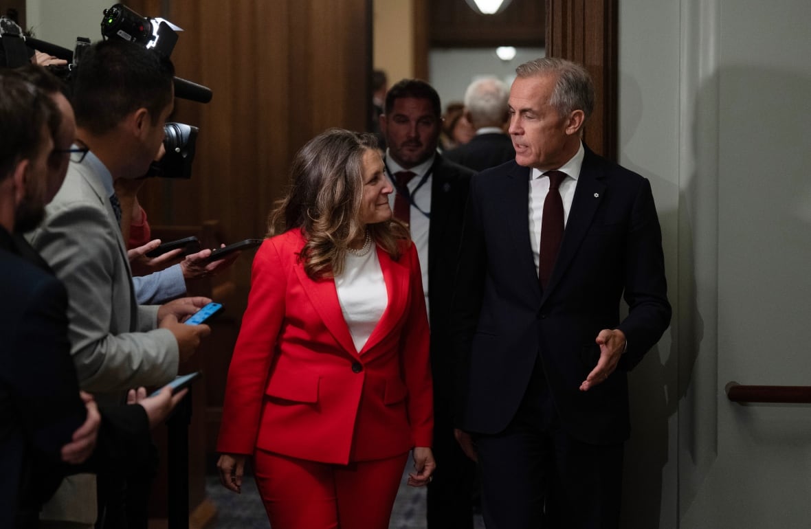 Two people speak while walking through a hall with reporters around them.