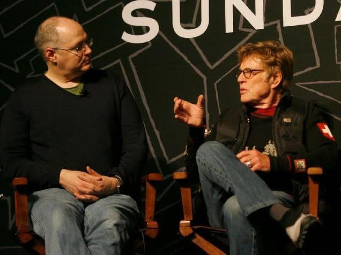 Two men sit next to each other in the directors of the directors, talking to each other. They both wear jeans and seem to be deeply involved in the conversation.