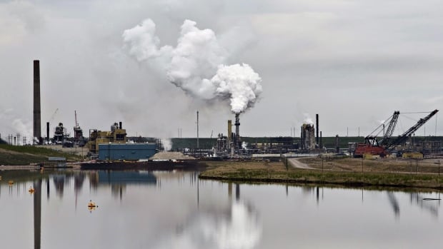 An industrial facility with smokestacks sending flumes in the air is reflected over water.