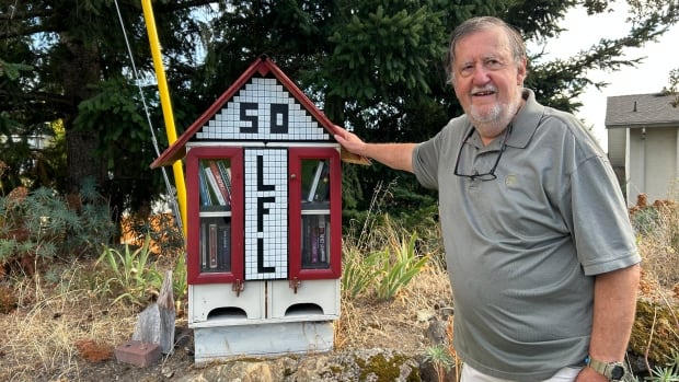 A senior man in a grey T-shirt and white bottoms stands next to a red and white little free library.