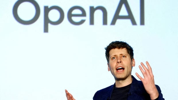 A man gestures while speaking. A large screen behind him says 'OpenAI'