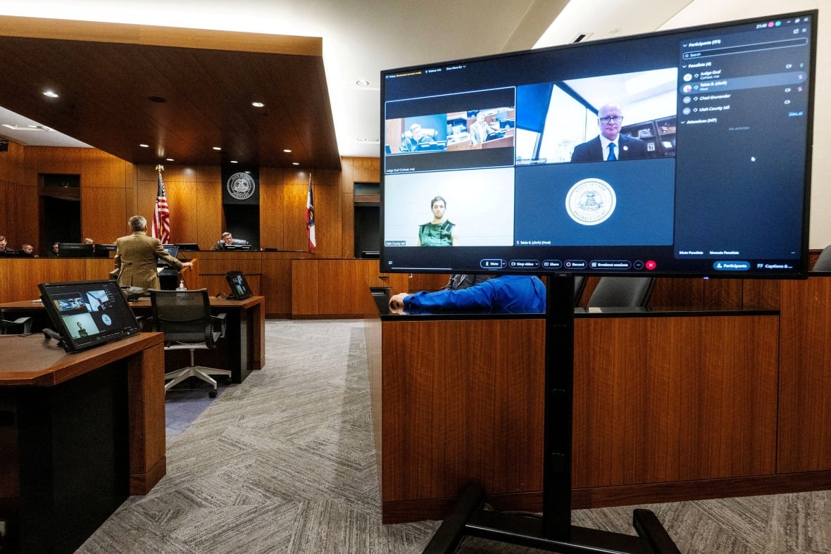 A wide shot of the court room, with a lawyer, sitting sitting and a large screen showing the defendant through the video link