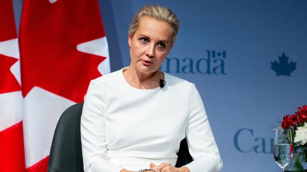 A blonde-haired woman with her hair tied back and wearing a knee-length skirt speaks while seated with her hands clasped in front of a Canadian flag.