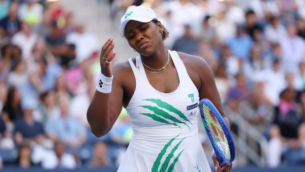A woman in a white tennis uniform reacts negatively during a match.