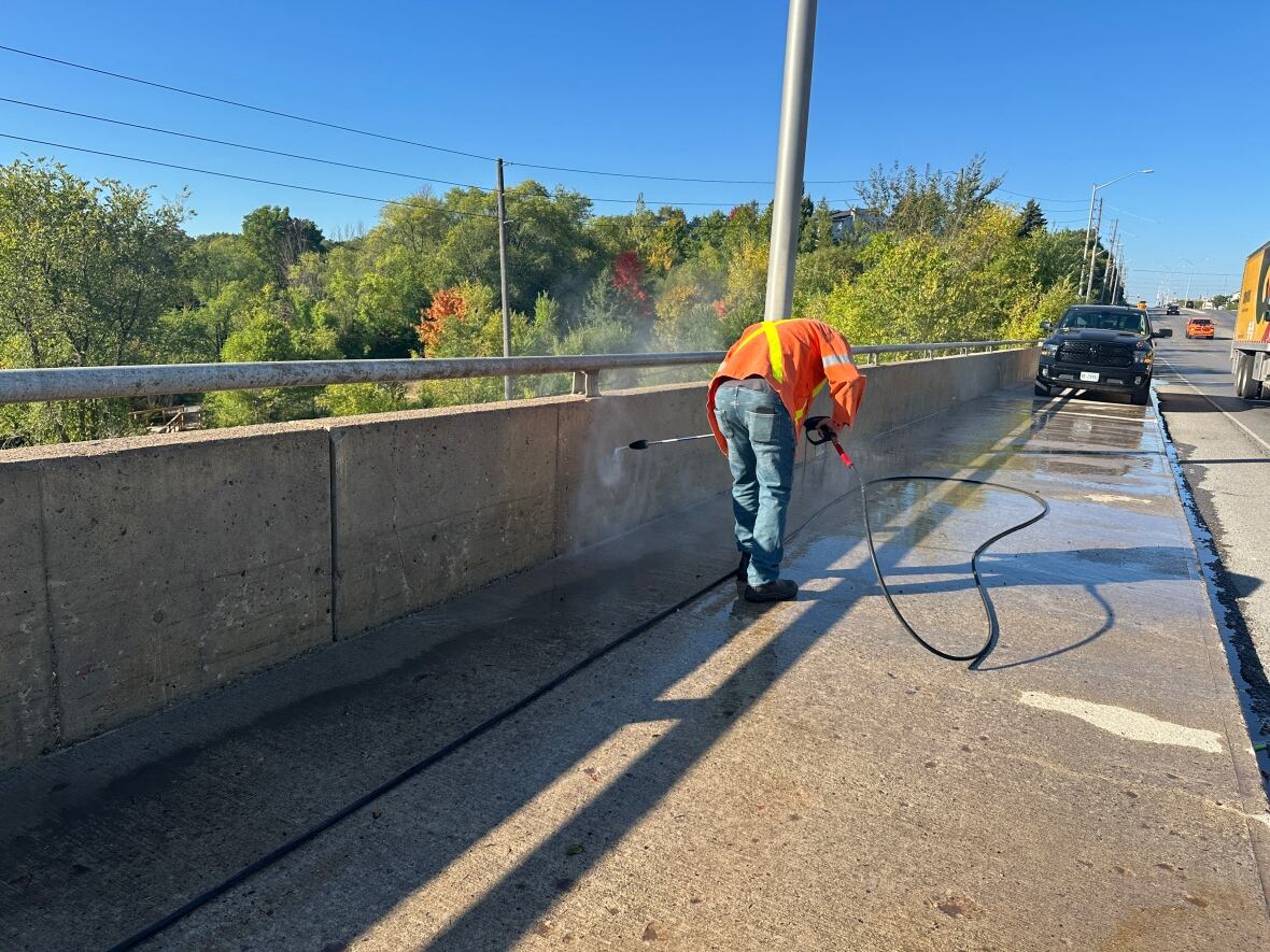 A man in a high visibility vest crouches near a concrete barrier overpass and sprays high-pressure water to remove graffiti. 