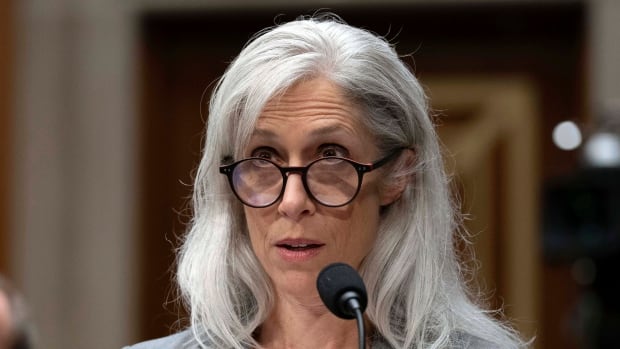 A greyhaired woman with her hair to her shoulders, wearing glasses and a blazer. 