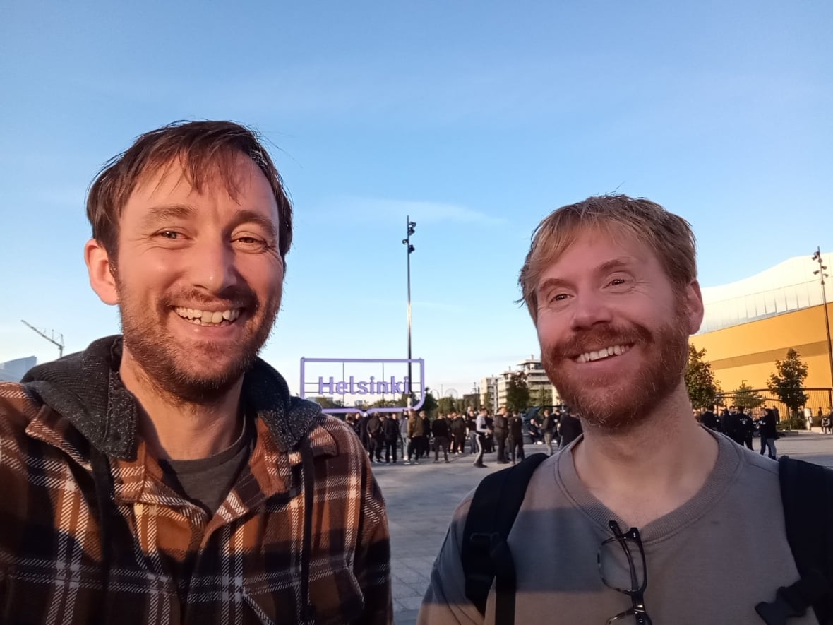 Dois homens estão na frente de uma grande placa lendo Helsinque