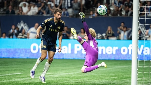 A soccer player in blue kit on left heads the ball past a diving goalkeeper wearing a mauve kit.