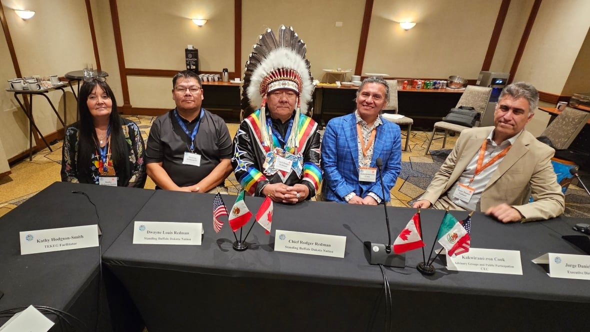 5 people, 4 men and 1 woman sit side by side at a long table, with one man in the middle wearing a feathered headdress and the women being on the end