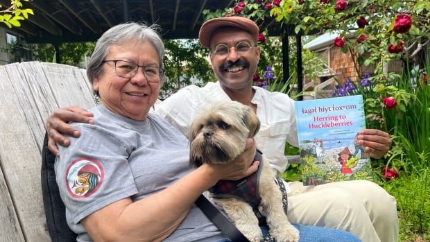 A woman holds a dog as she sits next to a man who has one arm around her and a book in the other hand