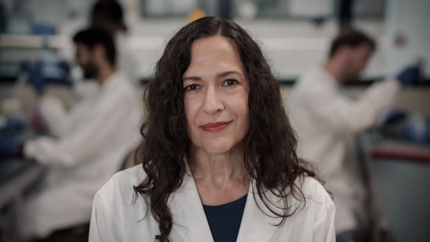 A woman with curly, brown hair past her shoulders wearing an embroidered lab coat stands with two scientists working at lab benches on both sides of her in the background.