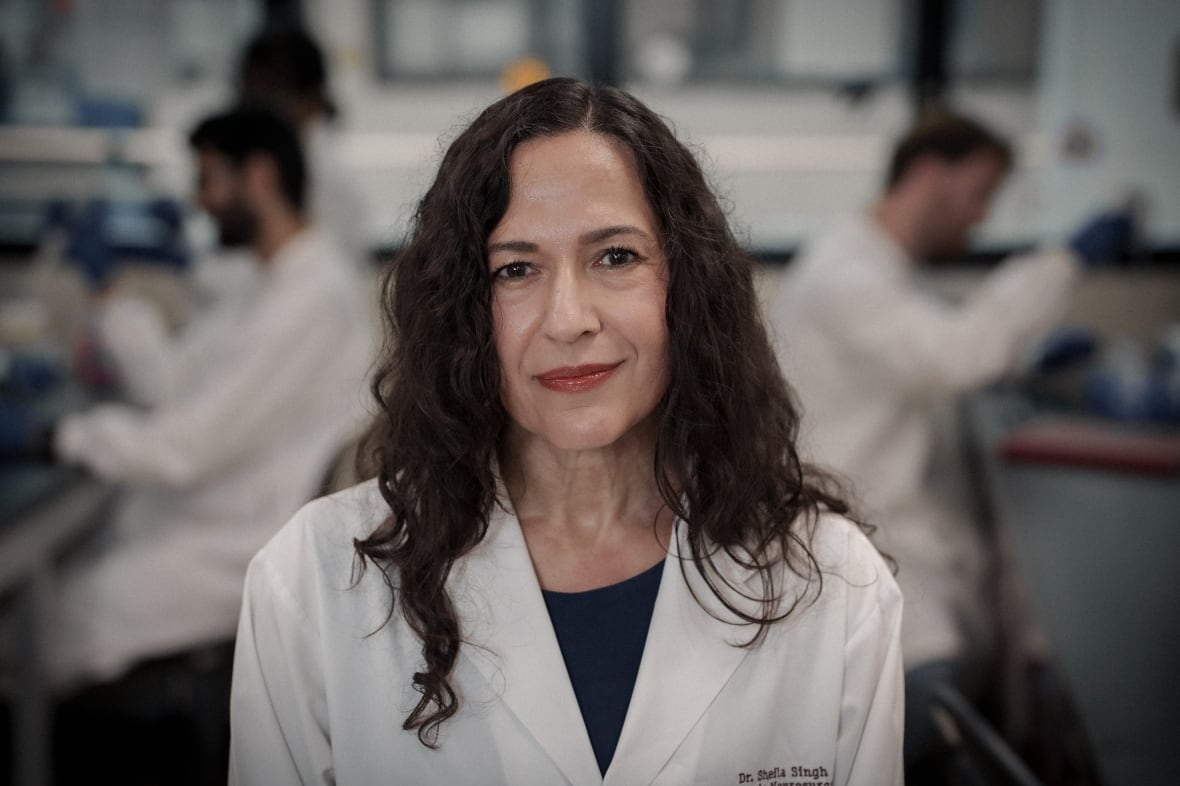 Una mujer con cabello castaño y rizado más allá de los hombros, vestida con una bata de laboratorio bordada, está de pie junto a dos científicos que trabajan en bancos de laboratorio a ambos lados de ella en el fondo.