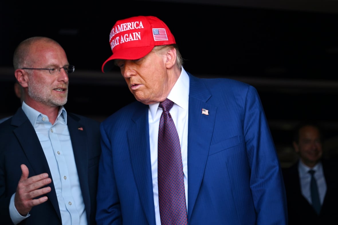 Een kalende man met een witte sikje praat met de Amerikaanse president Donald Trump, die een rode Make America Great Again-pet draagt.