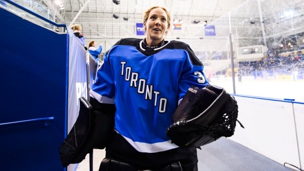 A women's hockey goalie walks on the outside of a rink, with her helmet off.