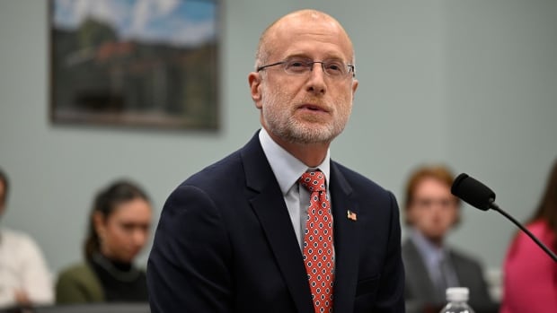 A balding bearded man with glasses wearing a dark suit and red dotted tie speaks while seated at a table.