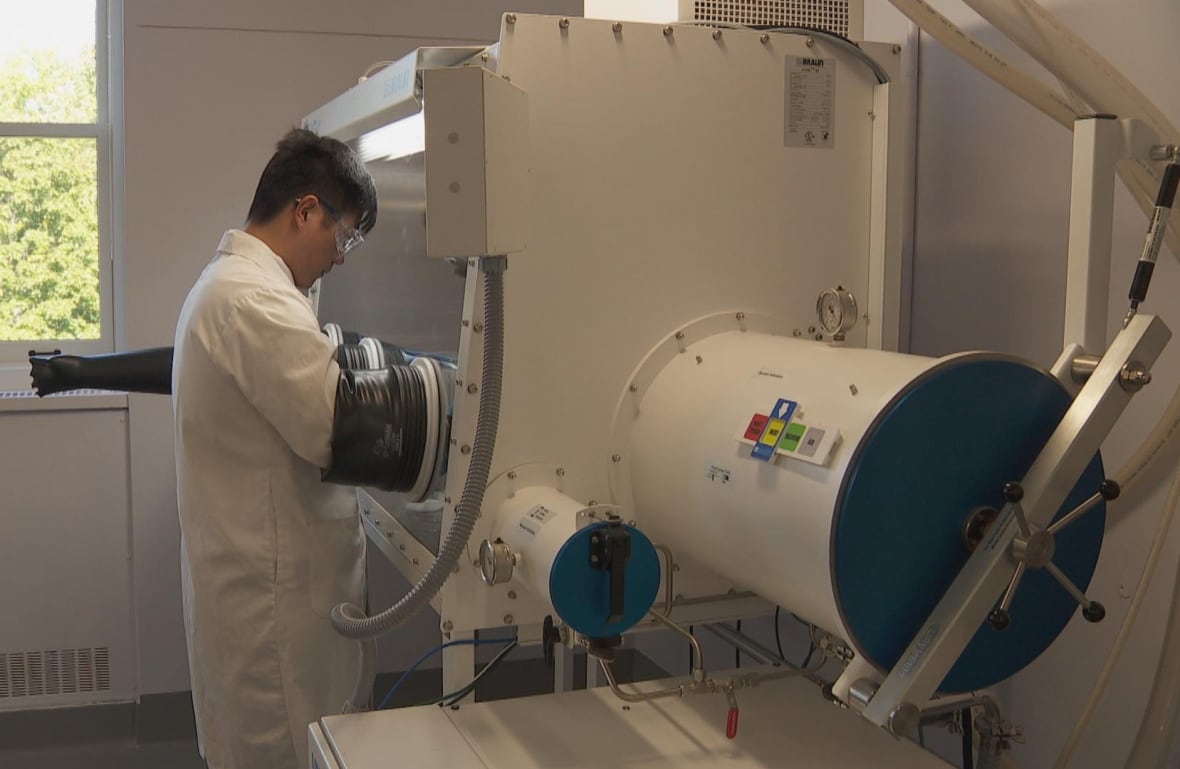Man working at a metal box like contraption roughly the size of a toddlers crib, with inverted rubber gloves attached to the box. Box is completely sealed to be air tight.