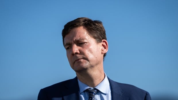 A close up image of Eby looking to the side, against a backdrop of the sea and mountains. He is wearing a blue suit, shirt and tie. 