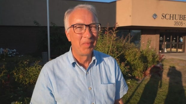 A man in a blue shirt and glasses stands outside in front of a community hall. 