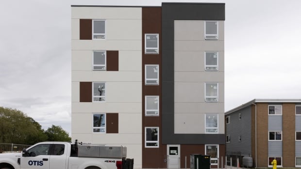 The brown and white square face of a five-story apartment building with windows faces the street. A white truck is parked out front