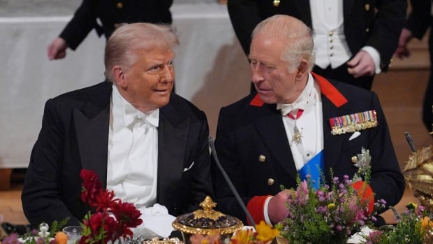 Two people formally dressed talk with one another while sitting at an elaborate dining table decorated with colourful flowers and including gold-plated dishes.