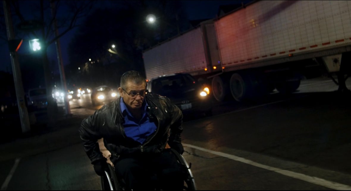 a person in a leather jacket uses a wheelchair on a road beside a transport truck