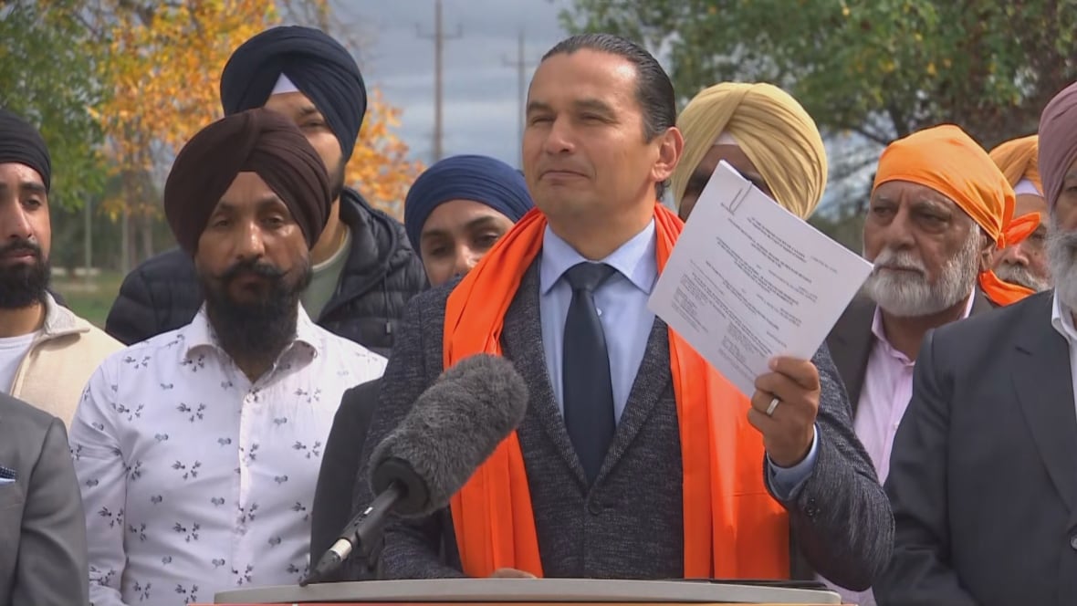 man wears suit and orange shawl, holding up a piece of paper at a podium