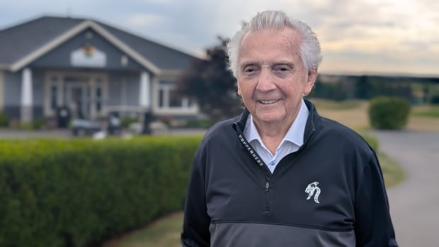 A man with white hair stands at the entrance to a golf course on a cloudy day.