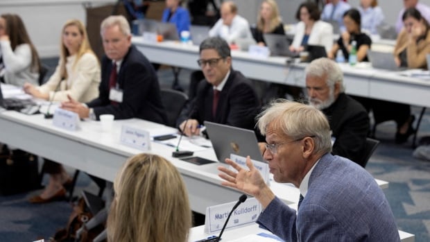 A man in a blue suit with glasses speaks around a table of experts in a boardroom