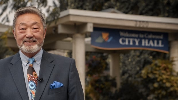 a man in a dark suit stands outside a building that has a sign reading "city hall"
