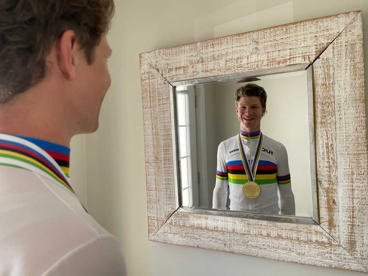 A young white man wearing a cycling outfit and a medal around his neck looks into a mirror on the wall. His reflection smiles back at him.