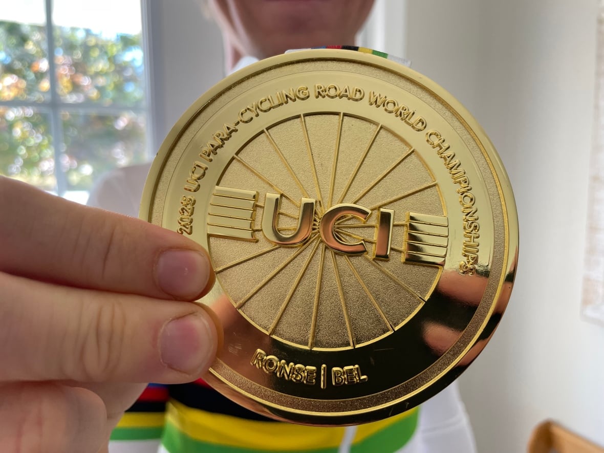 A close up on a gold medal that reads "2025 UCI Para-cycling Road World Championships" on the top and "Ronde, Belgium" on the bottom. It's being held by two fingers and you can see the person holding it slightly out of focus in the background.