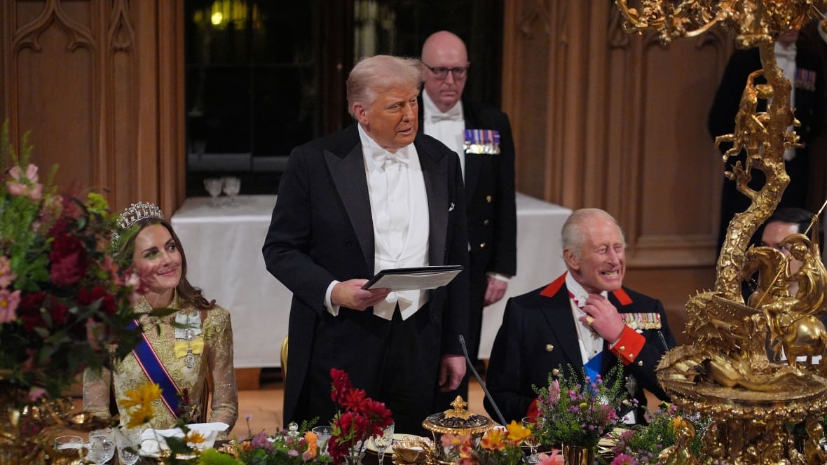 An elderly man stands while two people, a woman in a dress, scarf, crown and an elderly man who shines and adjusts his neck. They are on a long complex dining table during an official banquet.