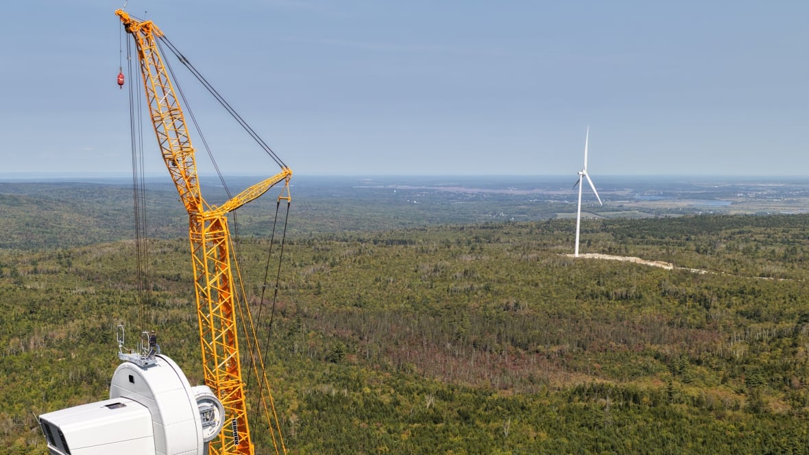 A worker stands at the top of a turbine hub as a crane lifts a blade into position to be installed at a wind farm. A completed turbine is seen in the background.